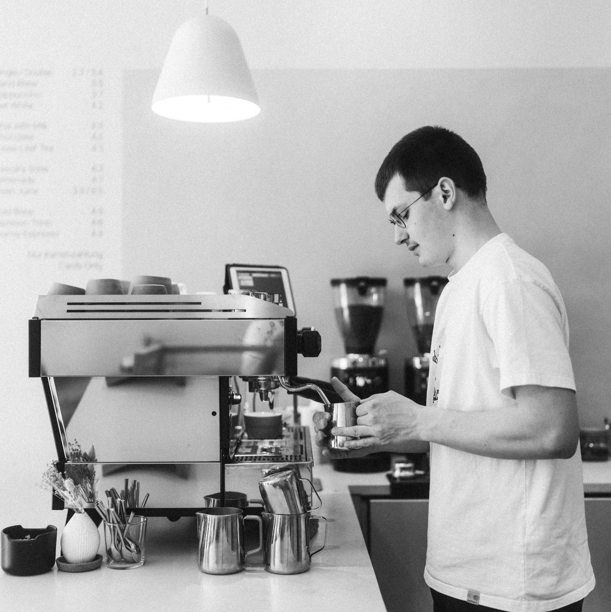 The barista in standing behind an espresso machine by la Marocco and is currently steaming the milk with the steam wand. The picture is black and white.