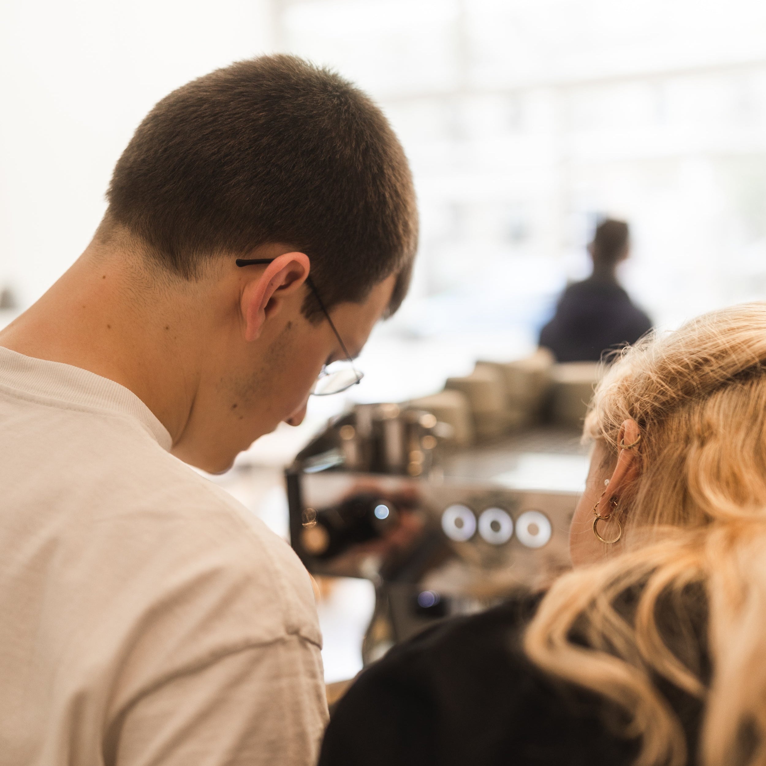A male barista is pouring latte art (not currently visible in the frame) while another female barista obeserves the pouring of the latte art.