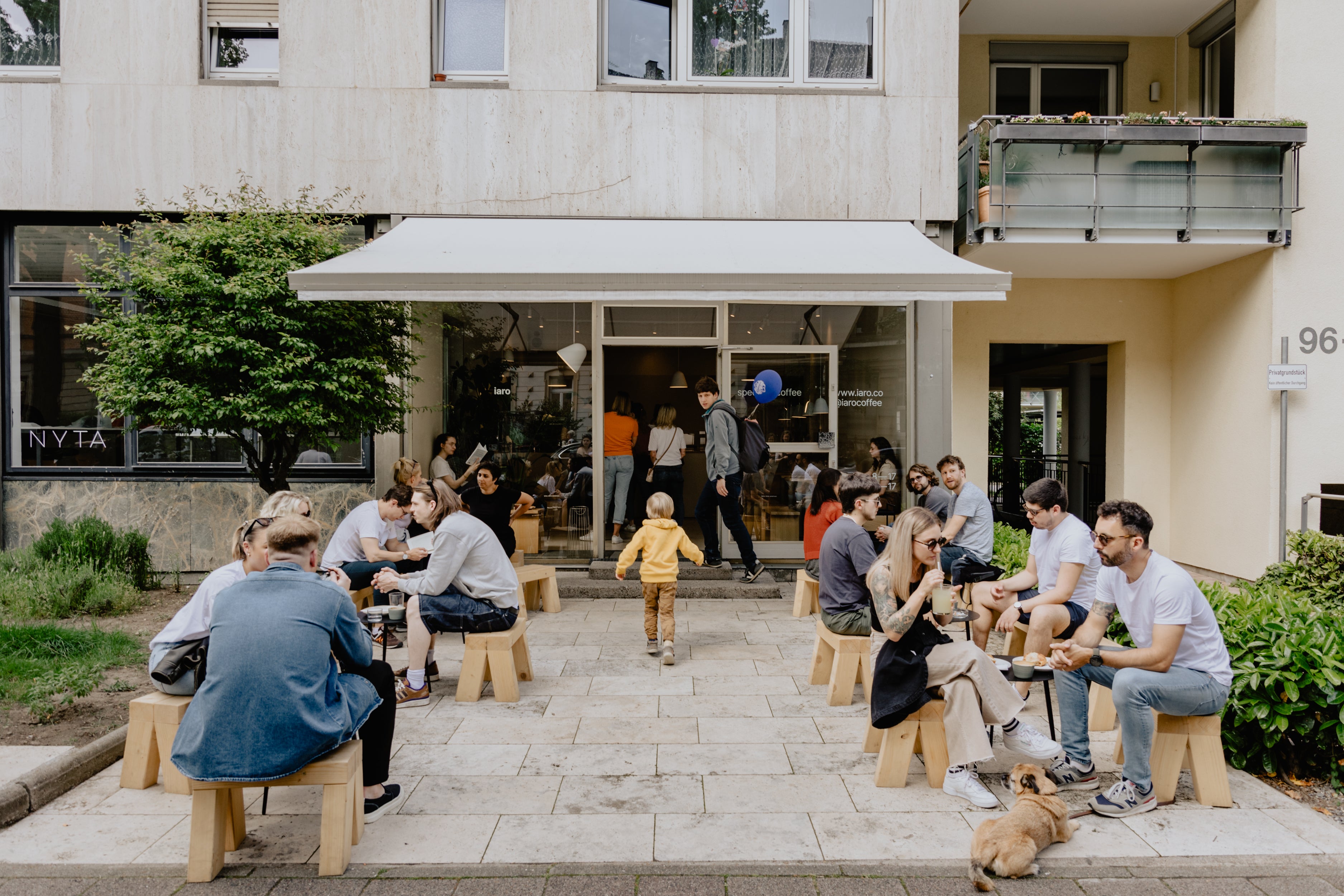 A lot of people are sitting on wooden benches and stools on the terrace of the coffeeshop iaro in the Weststadt in Karlsruhe on a cloudy summer day.