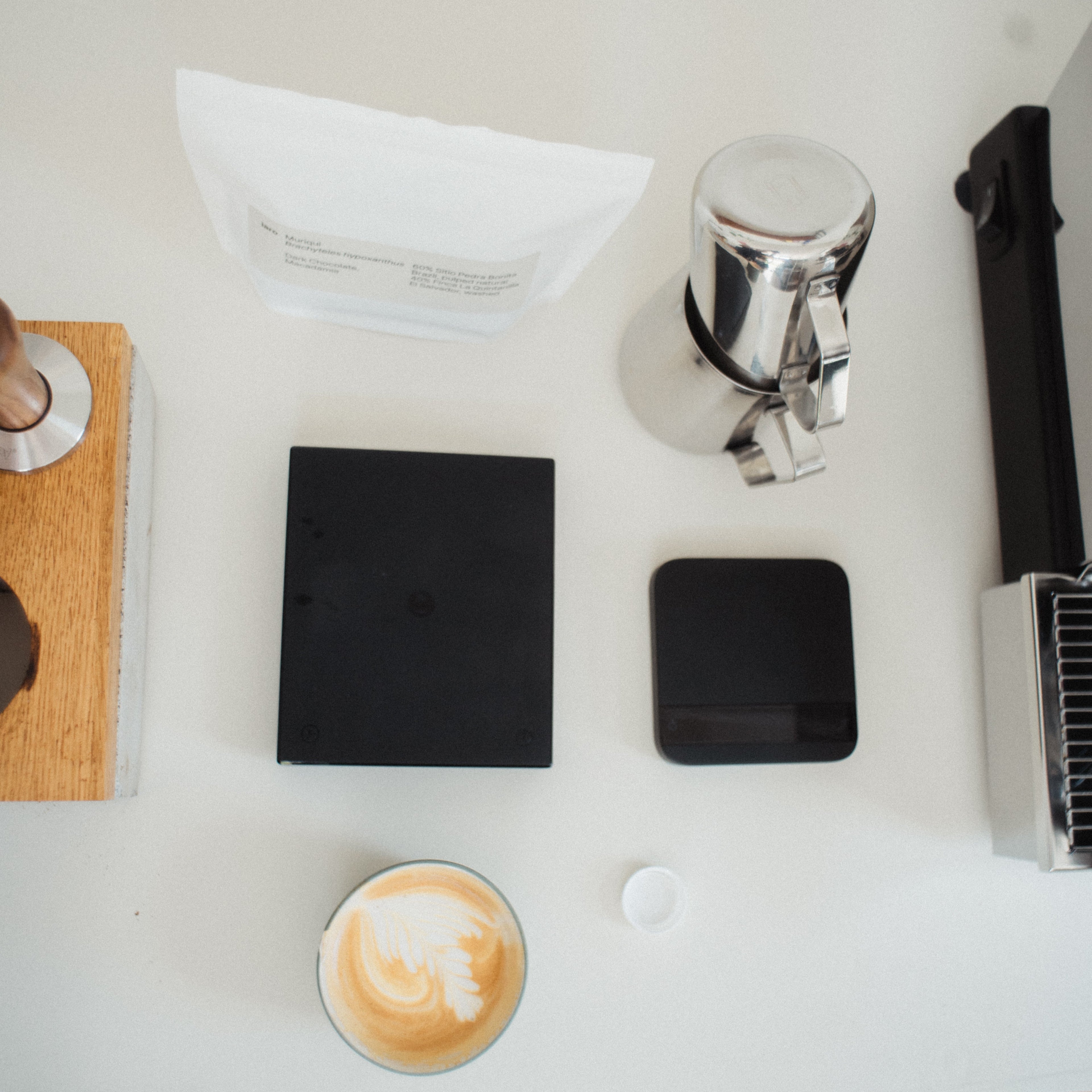 The essential home barista tools are layed out on a white tabletop next to a coffeemachine.