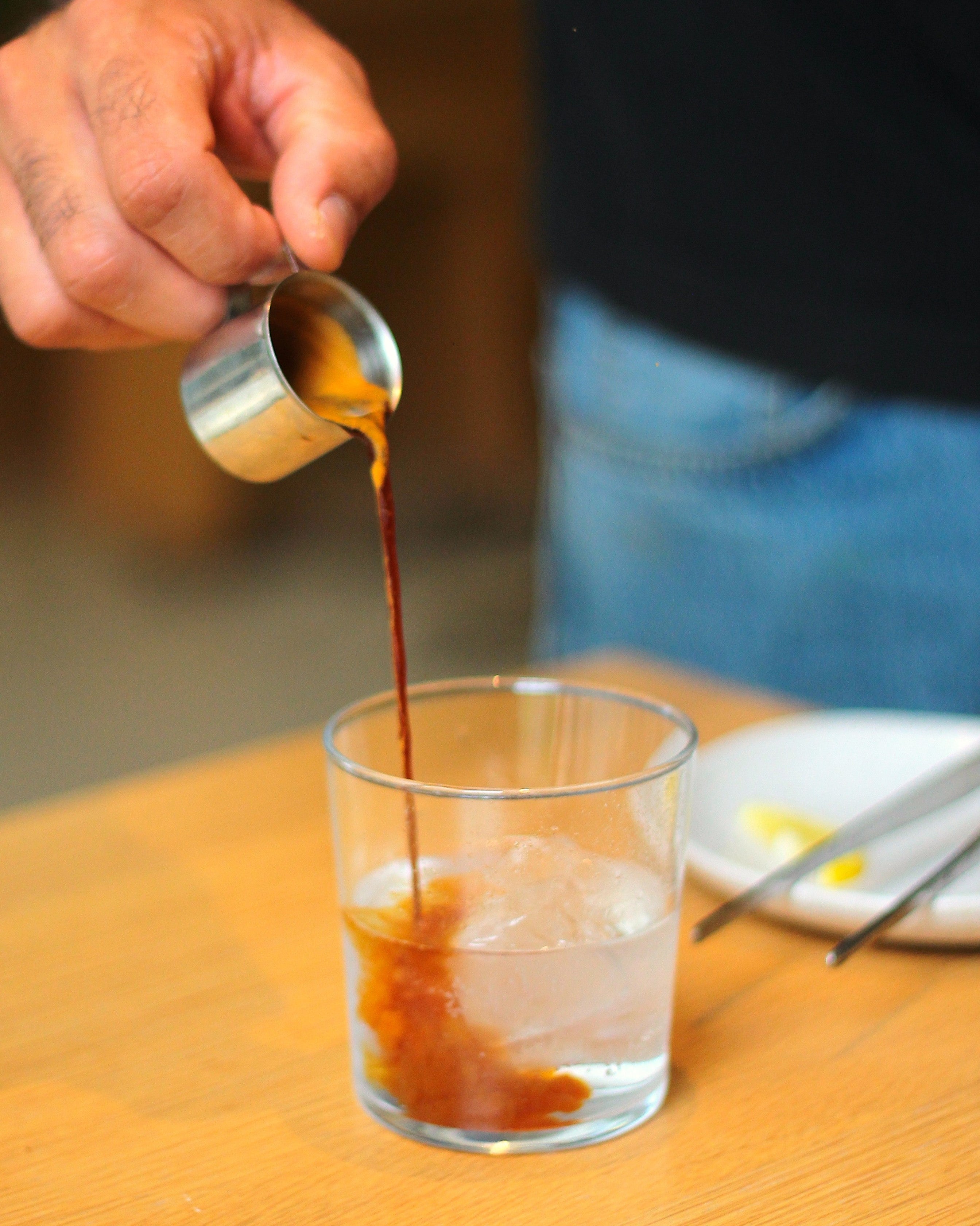 A sho of espresso is being poured from a small stainless jug into a glass filled with icecubes and tonic water.