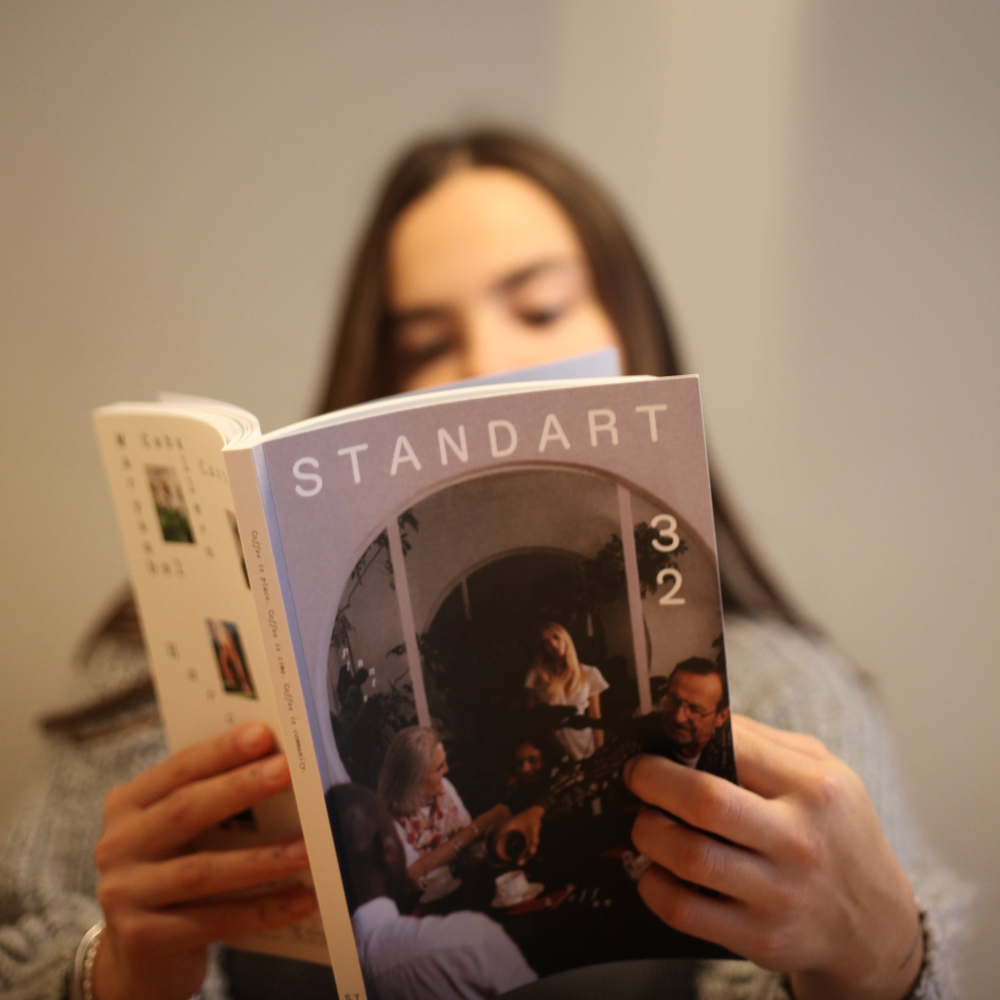 A girl is holding up the 32nd issue of the Standart coffee mag with both hands and reads. Her face if half-covered by the magazine and can be seen, blurred out in the backround