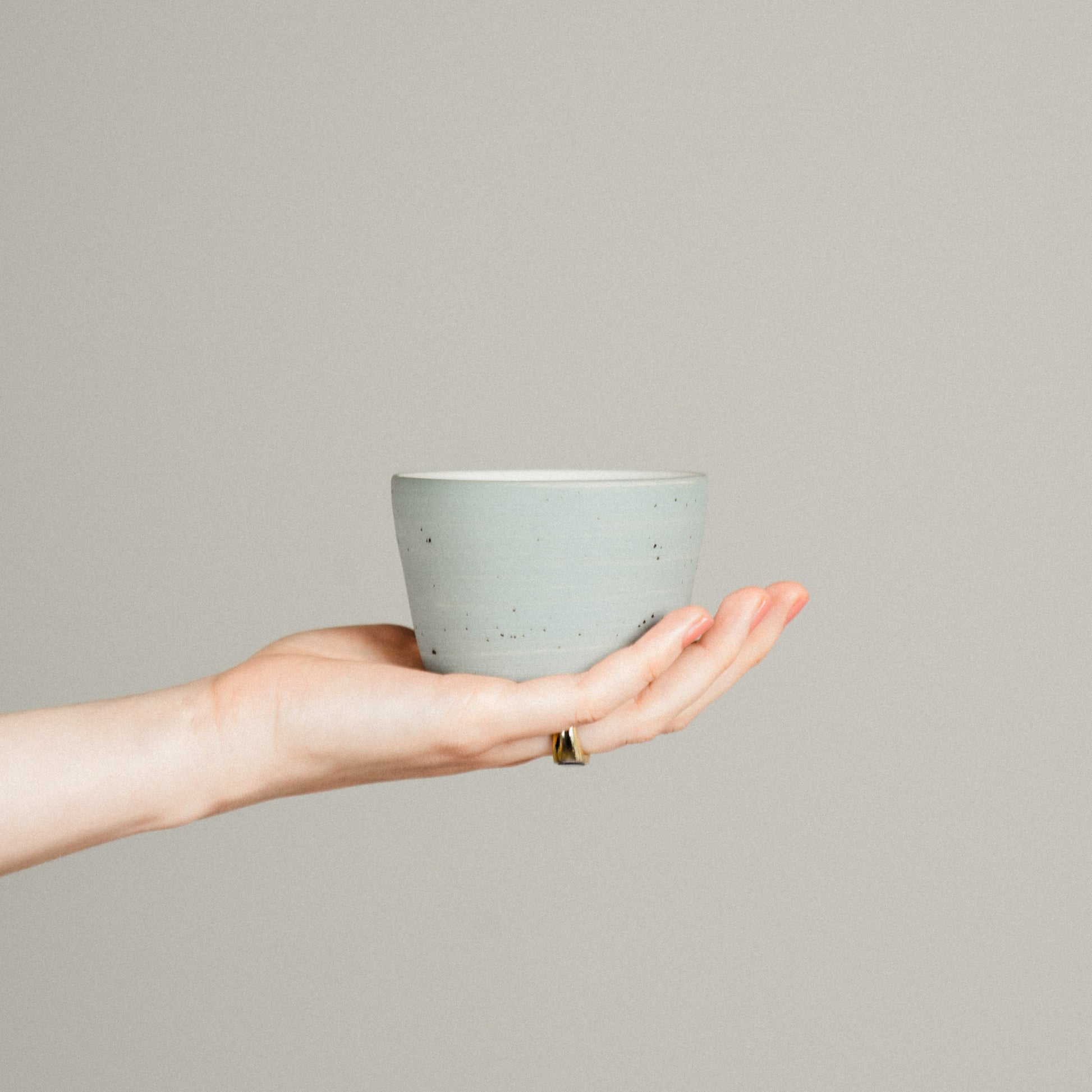 A delicate hand is holding a specialty coffee cup on its palm in front of a neutral backround.