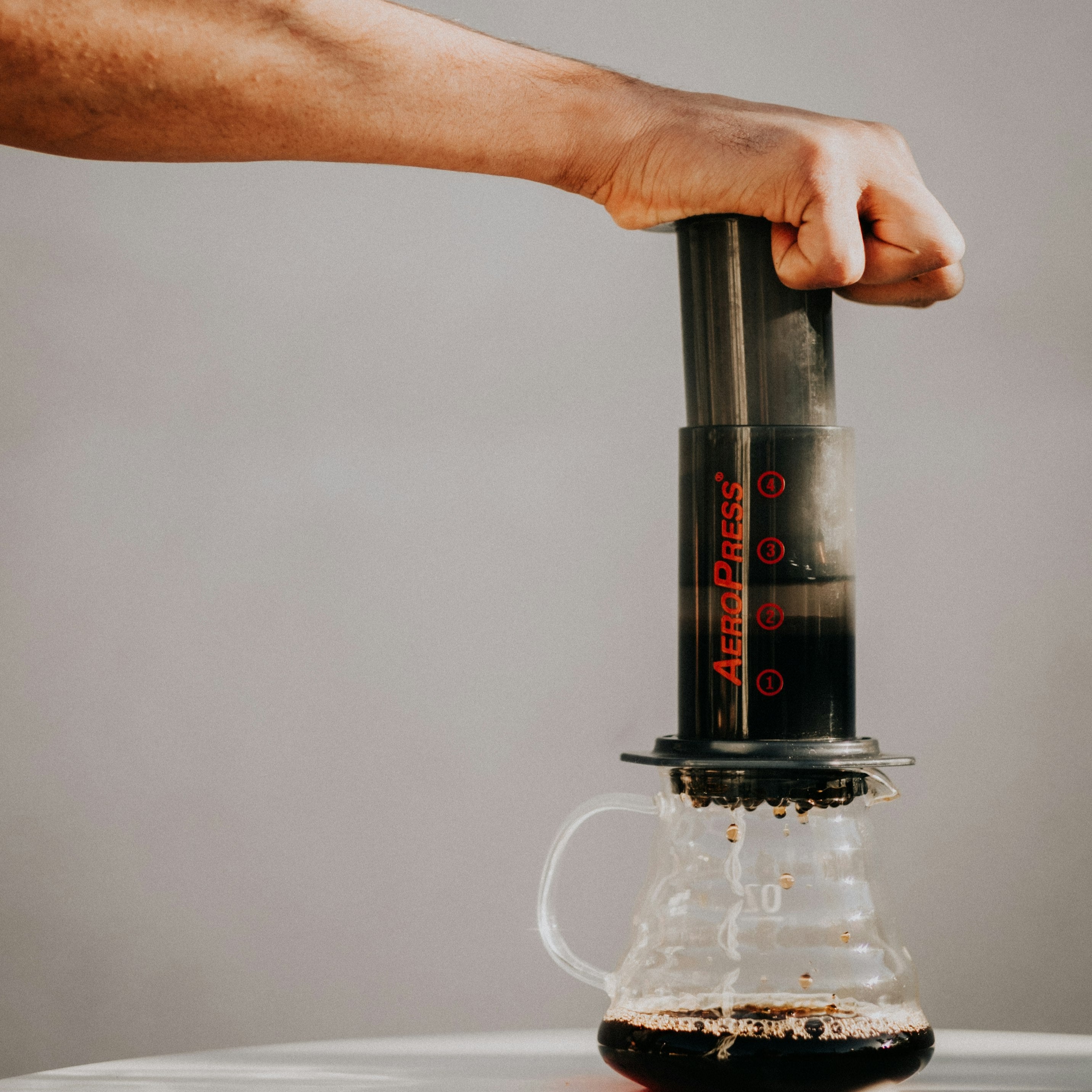 A hand presses down on an AeroPress Coffeebrewer to release the coffee into the glass jug beneath