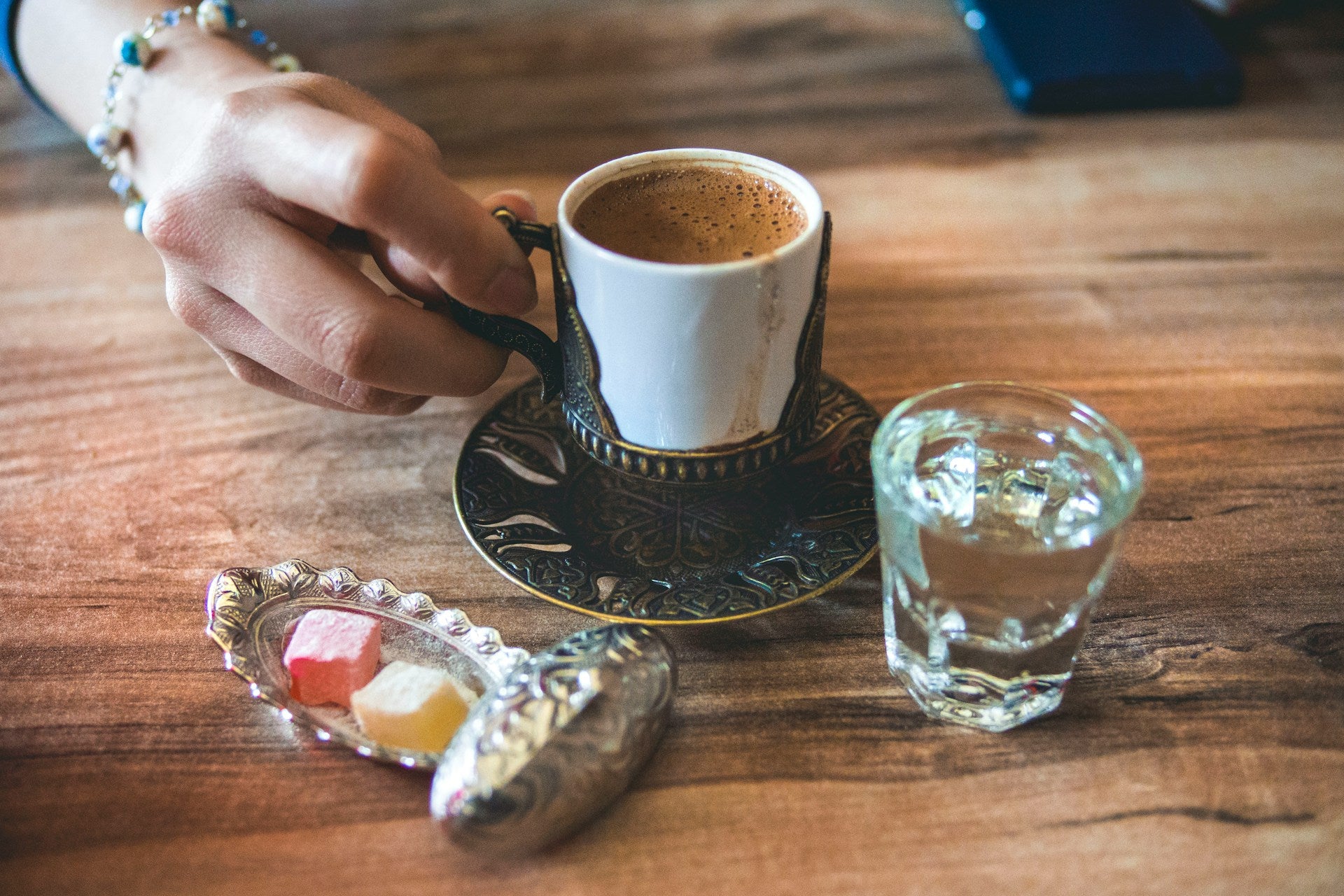 A delicate hand is reaching for a small cup framed in metal. The cup is filled with coffee and is standing next to a tiny glass of water and a silver tray with turkish delights.