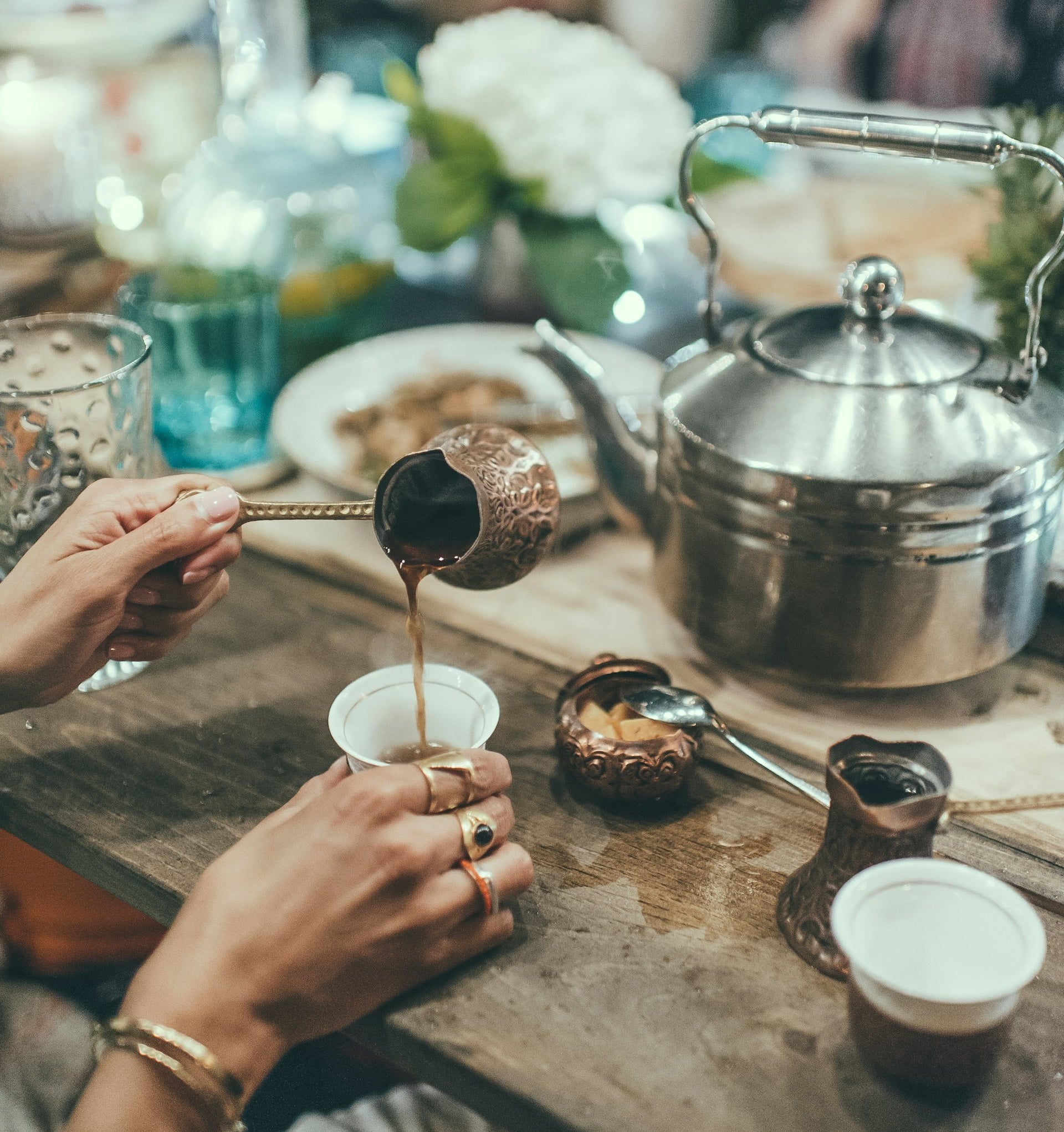 Delicate hands with golden jewelery pouring coffee from cezve into a small traditional ceramic cup. The backround is blurred by bokeh and filled with a big teapot and other items.