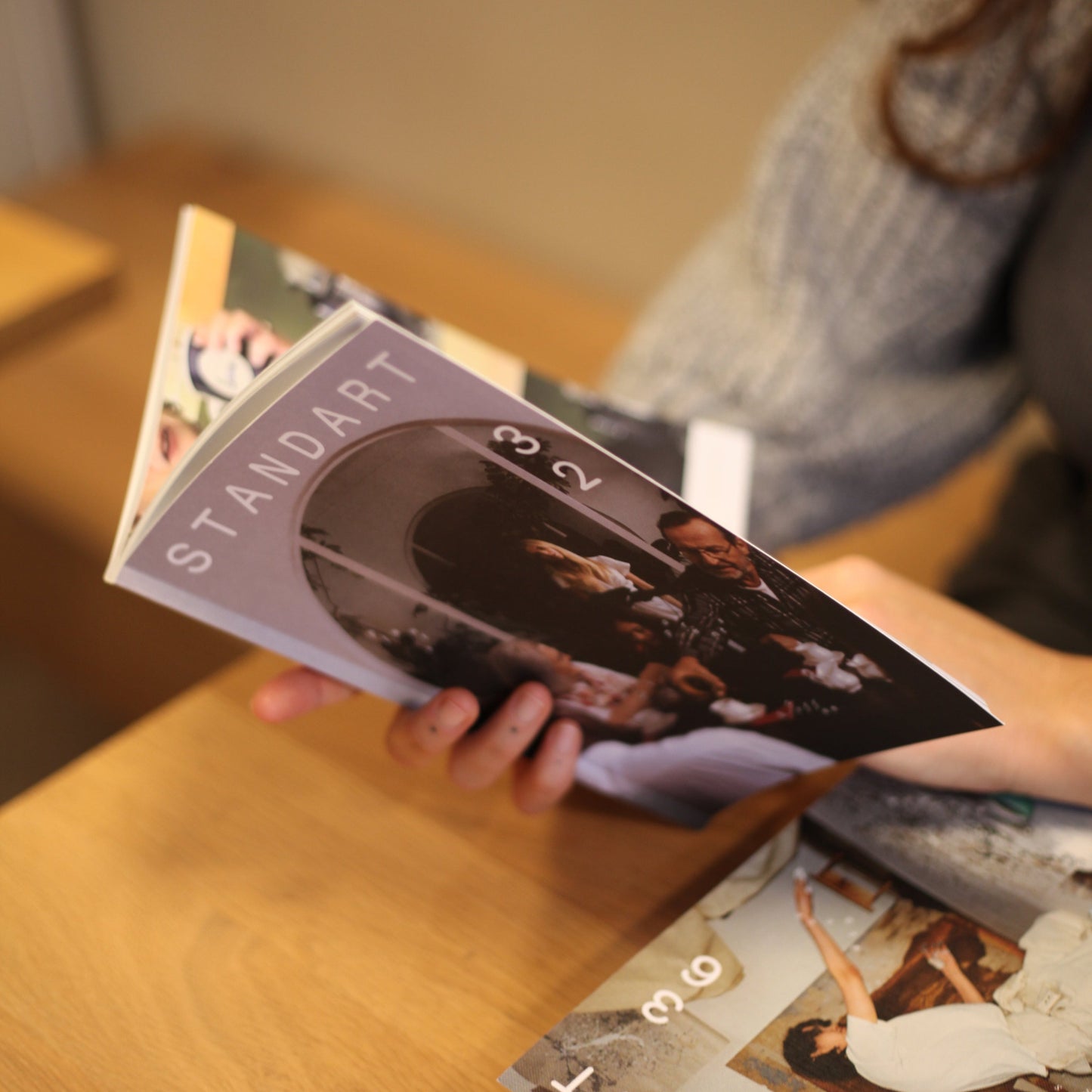 A woman is sitting at a table in a cozy environment and looking at an opened Standart magazine she is hlding in her hand.