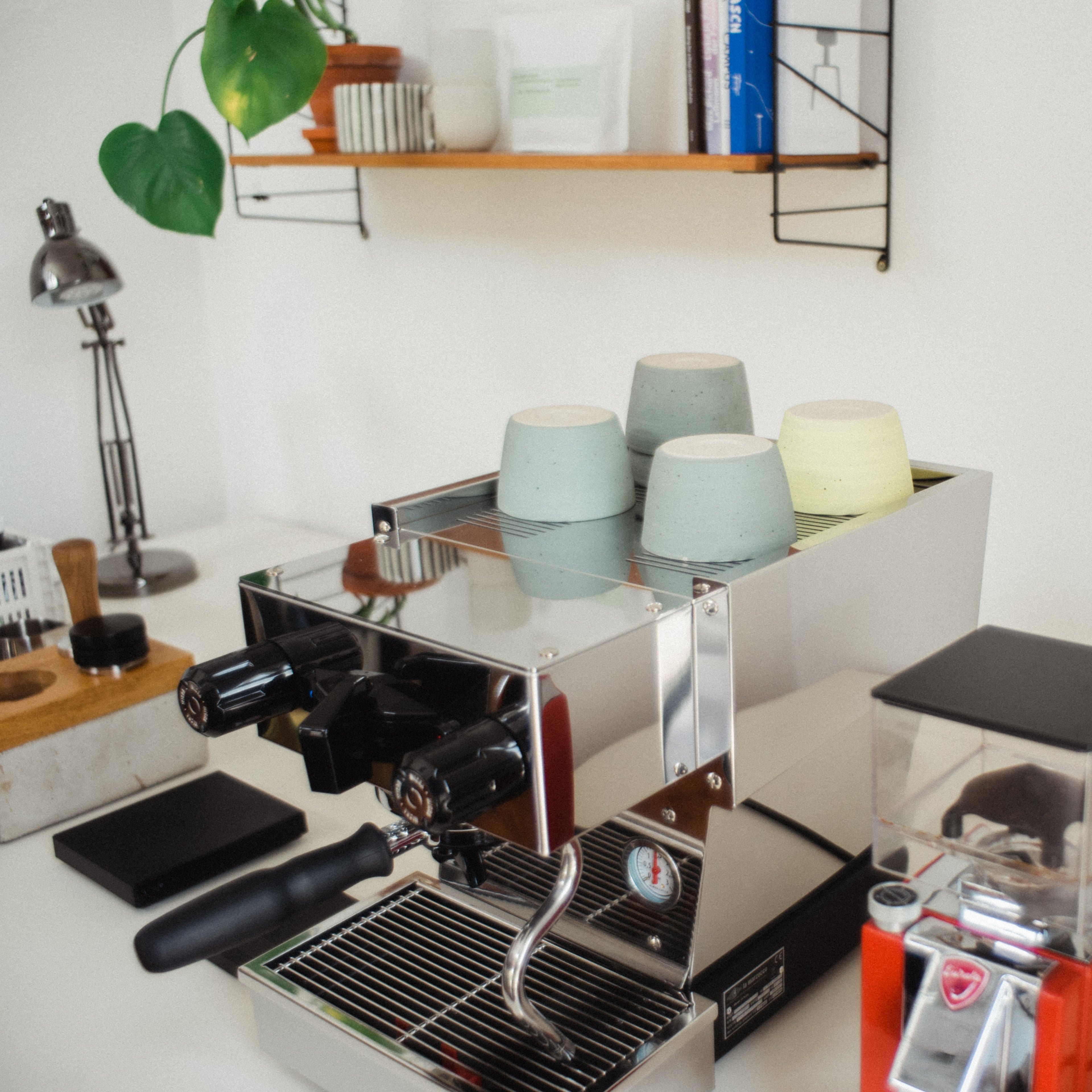 A la Marzocco Linea Micra is standing on a table next to a coffee scale as well a red Eureka grinder. A couple of iaro specialty mugs are placed on top of the machine.