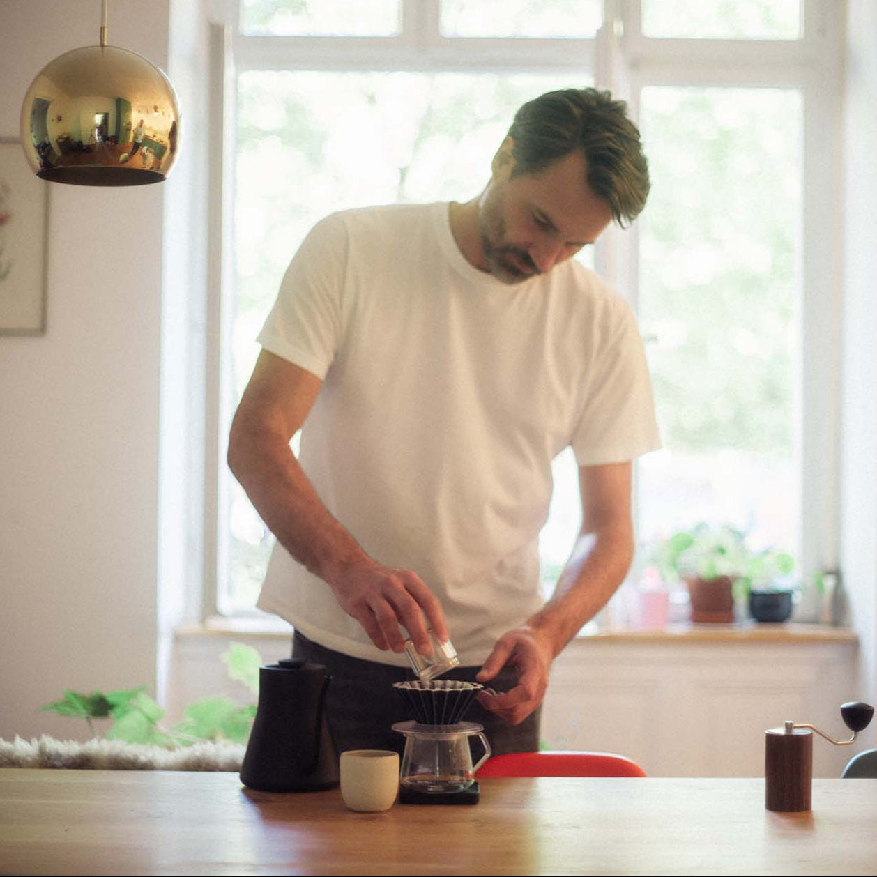 A man in a white t-shirt is pouring ground coffee into an origami coffee filter from the glass collection cup of his hand grinder.