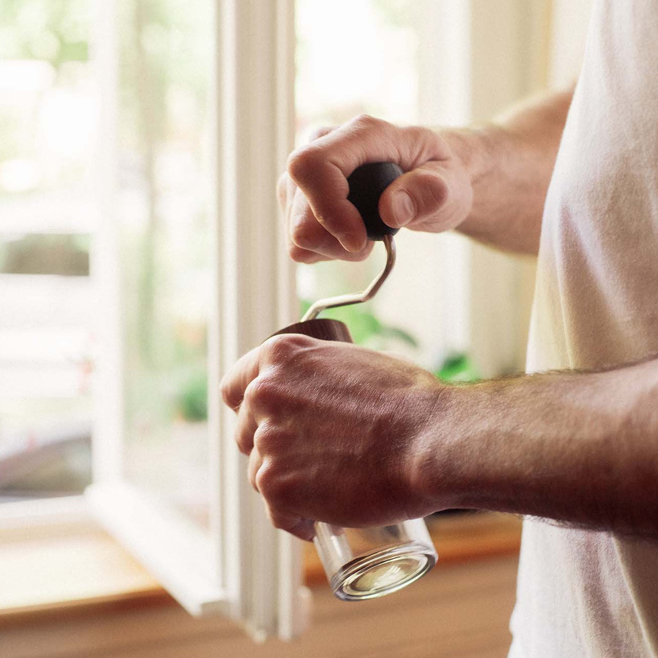 A man is holding the body of a coffee hand grinder firmly with one hand while turning the handle of the grinder with the other.