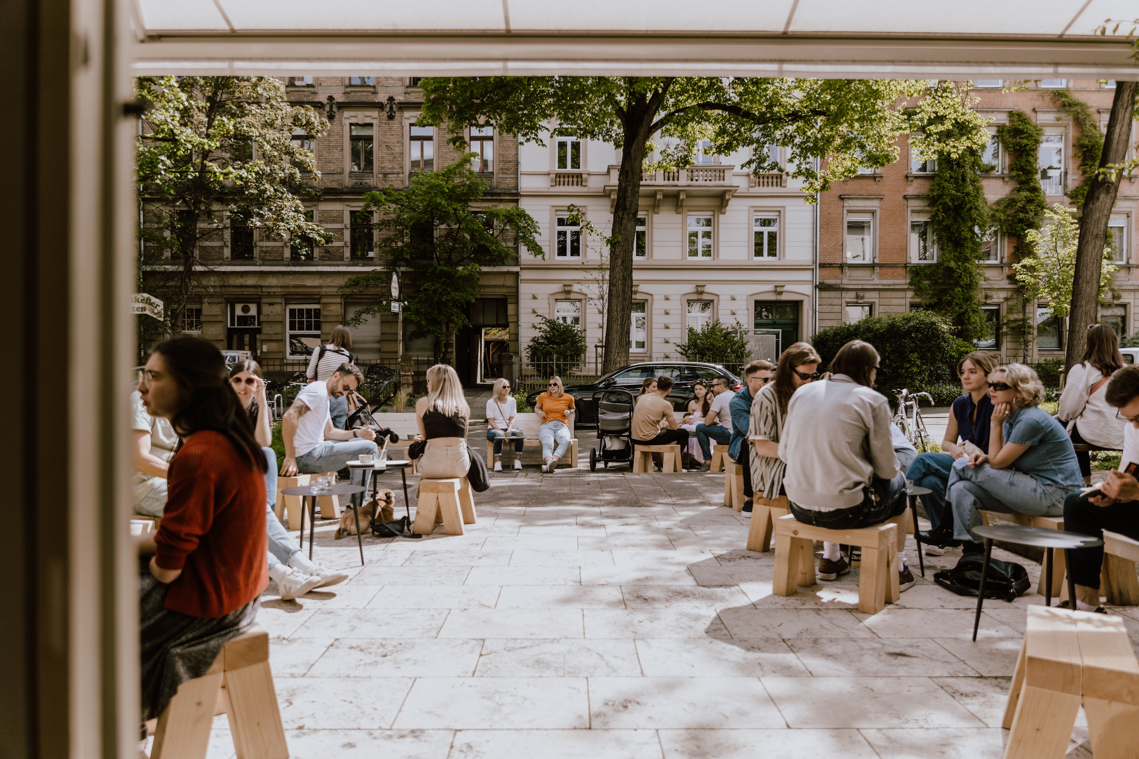 The terrace of a specialty coffeeshop on a busy and sunny day. It shows the view from the inside of the cafe with the trees and houses on the opposite side of the street, as well as the terrace packed full of people drinking coffee and enjoying the sun.