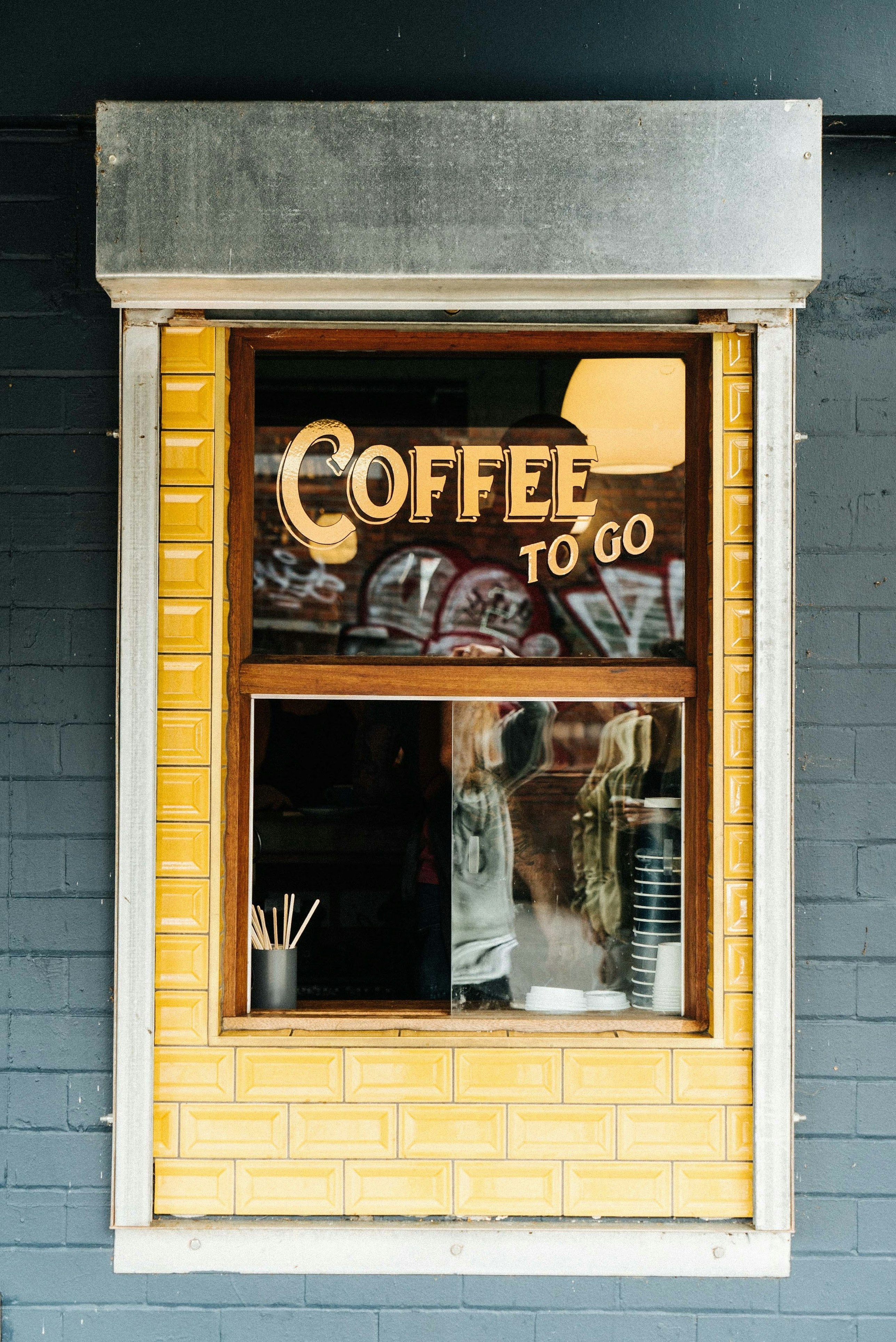 Window with antique golden font spelling "coffee to go". The wall surrounding the window is made up of yellow tiles. The window is open and meant to sell and serve coffee to go.