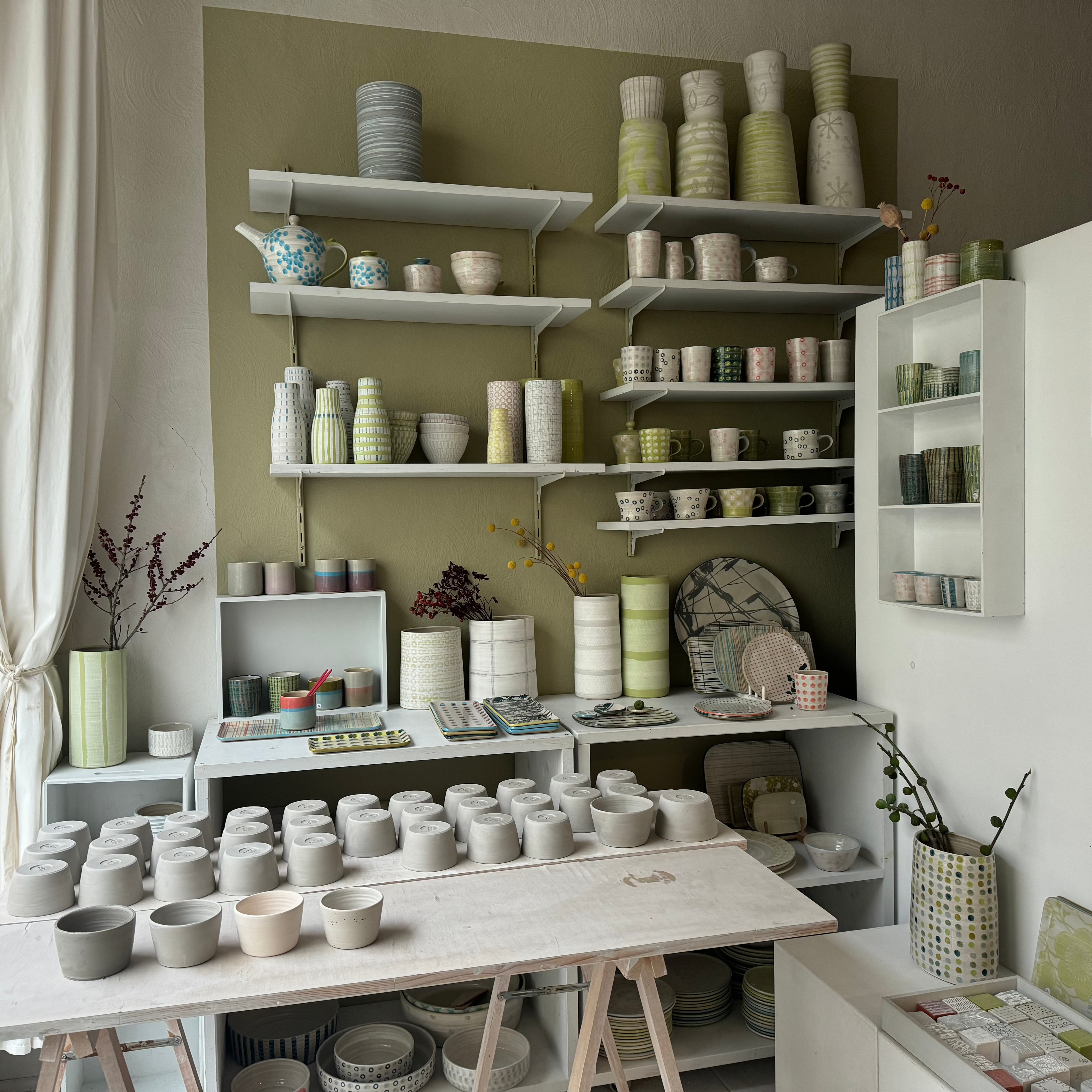 The atelier of artist Jutta Becker. A lot of raw iaro mugs are layed out on the workbench to dry. In the backround a bunch of different pieces by are displayed in tall wallmounted shelves.