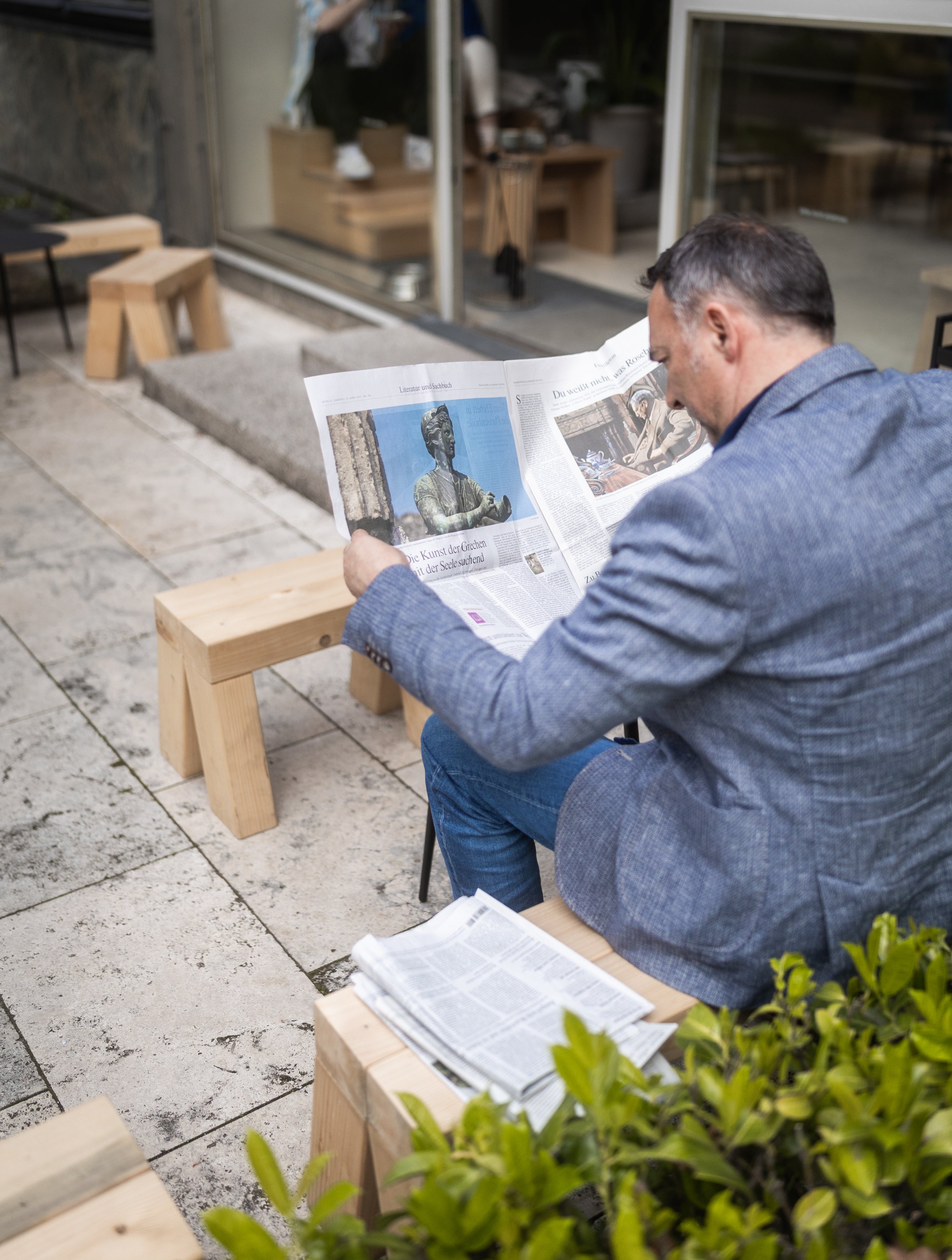 Man wearing a light blue blazer sitting on a wooden bench reading a newspaper in front of a coffeeshop