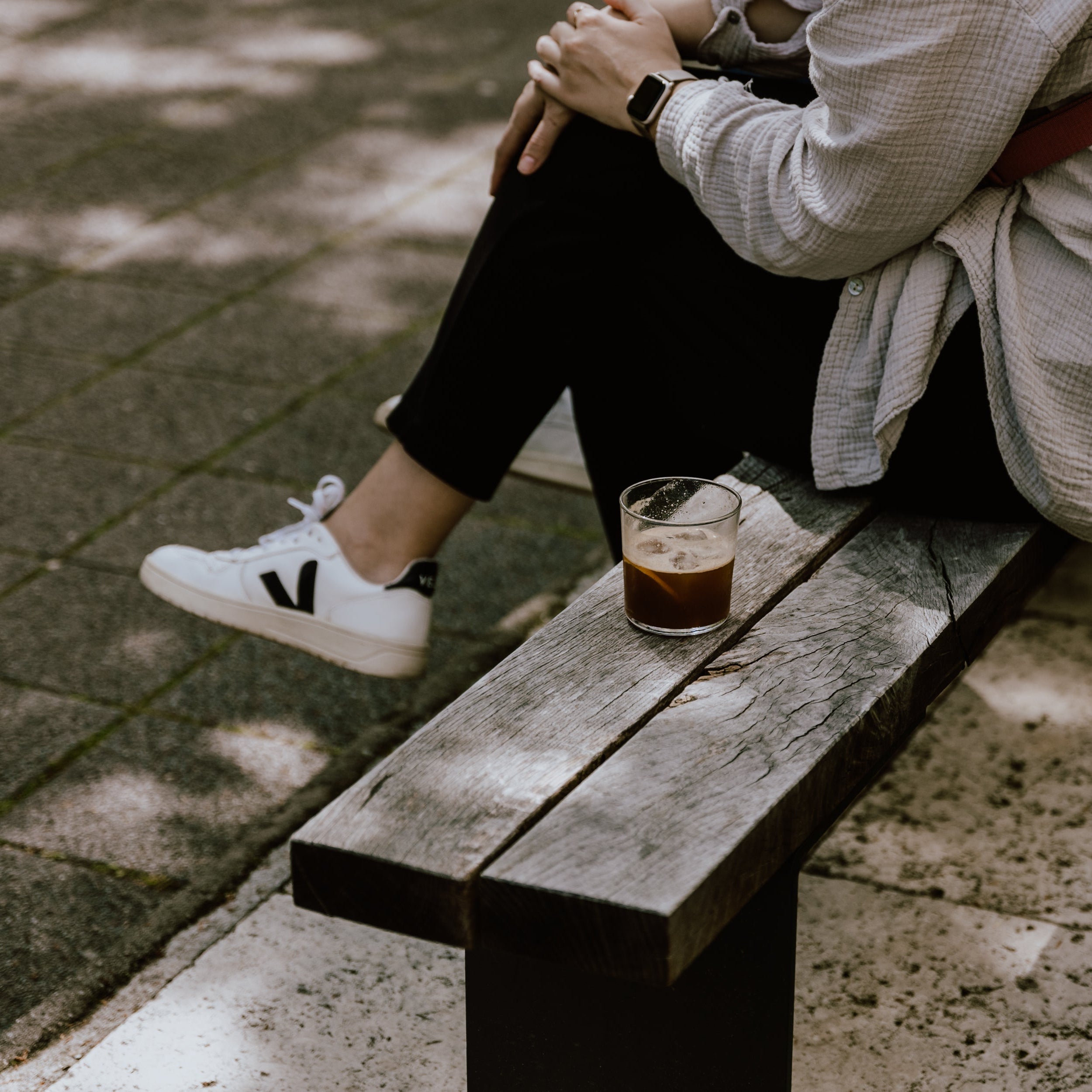 A glass filled with espresso tonic next to a casually dressed person, sitting on a bench.