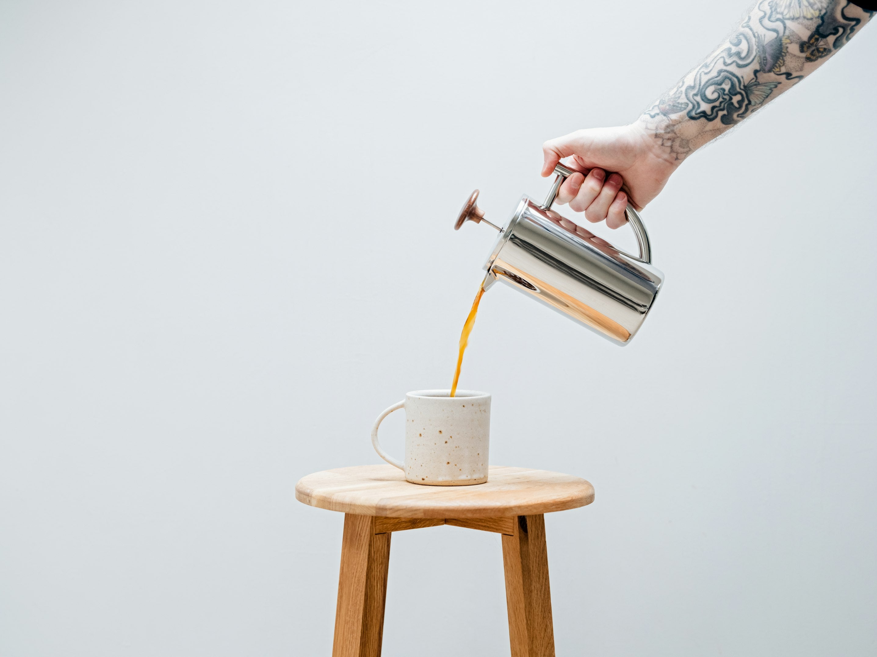 A forearm reaching into the frame of the picture pours coffee from a shiny metal french press coffee maker into a beige mug standing on a wooden stool