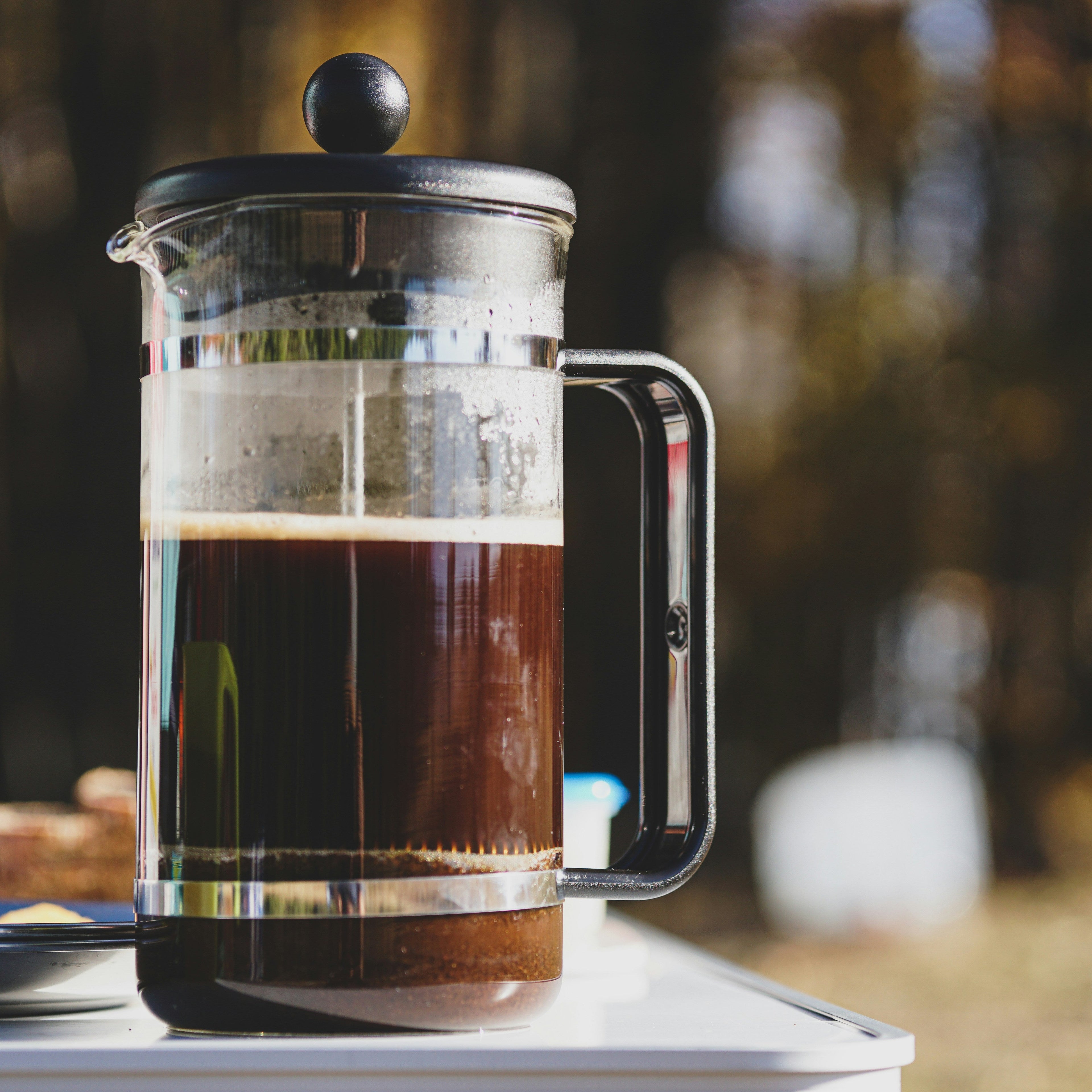 A close up of a french press caraffe with freshly brewed coffee inside. The caraffe is standing on a white camping table in the woods.