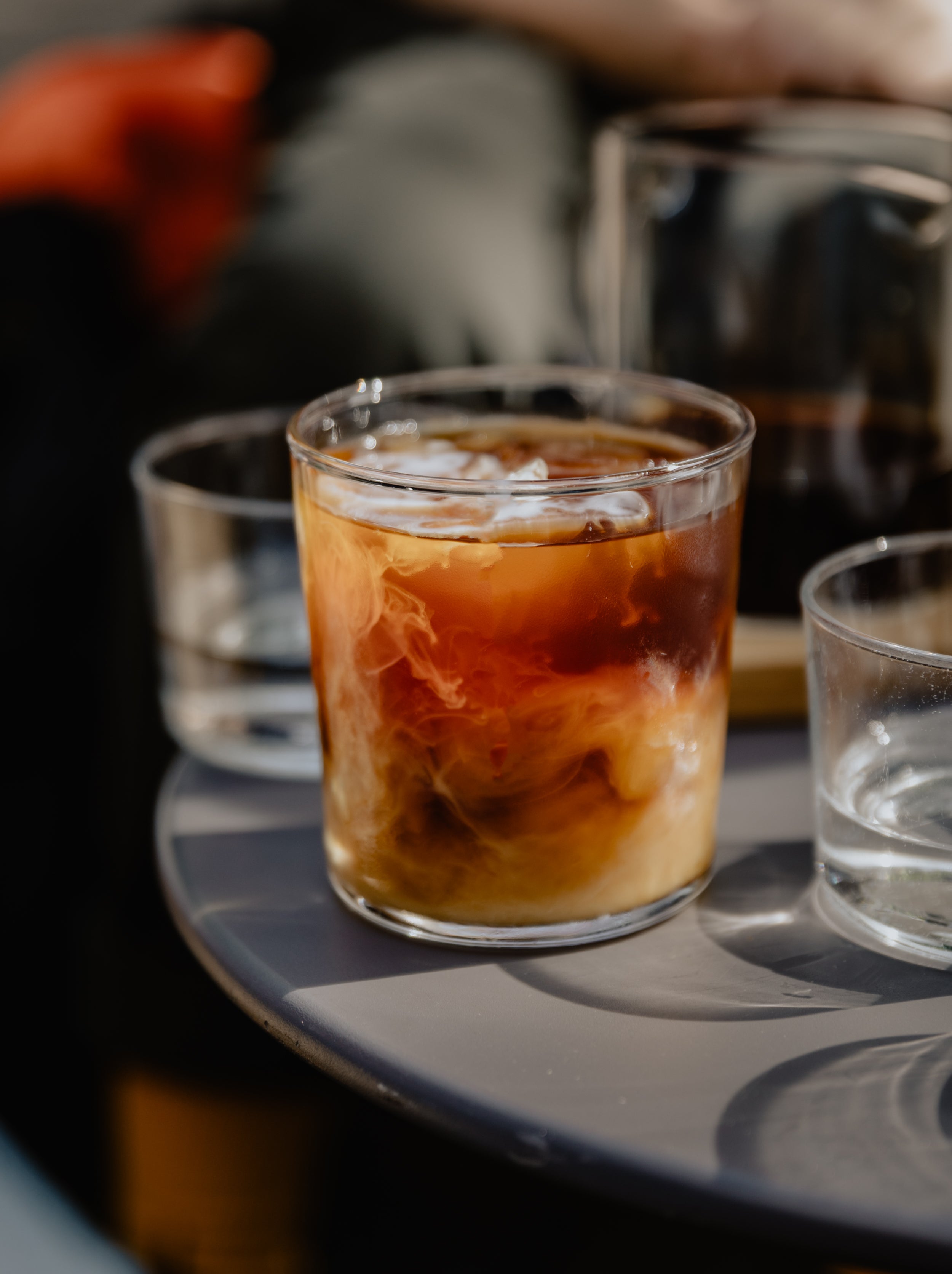 A glass of cold brew coffee with a bit of milk just poured into the drink resulting in a cloud-like pattern forming. It stands in between other glasses filled with water.