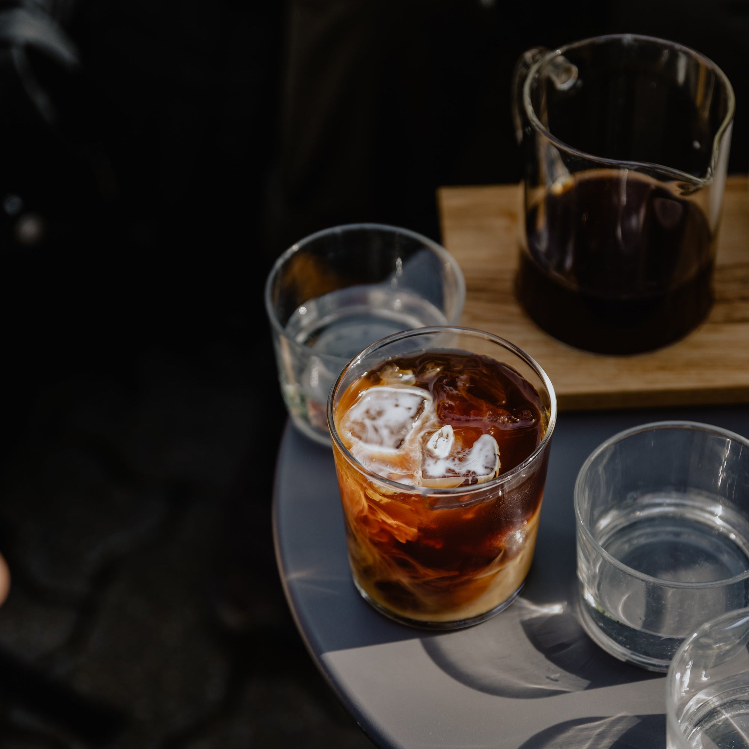 A glass of cold brew coffee with a bit of milk standing on a small grey table with several water glasses and a caraffe of hand brew filter coffee