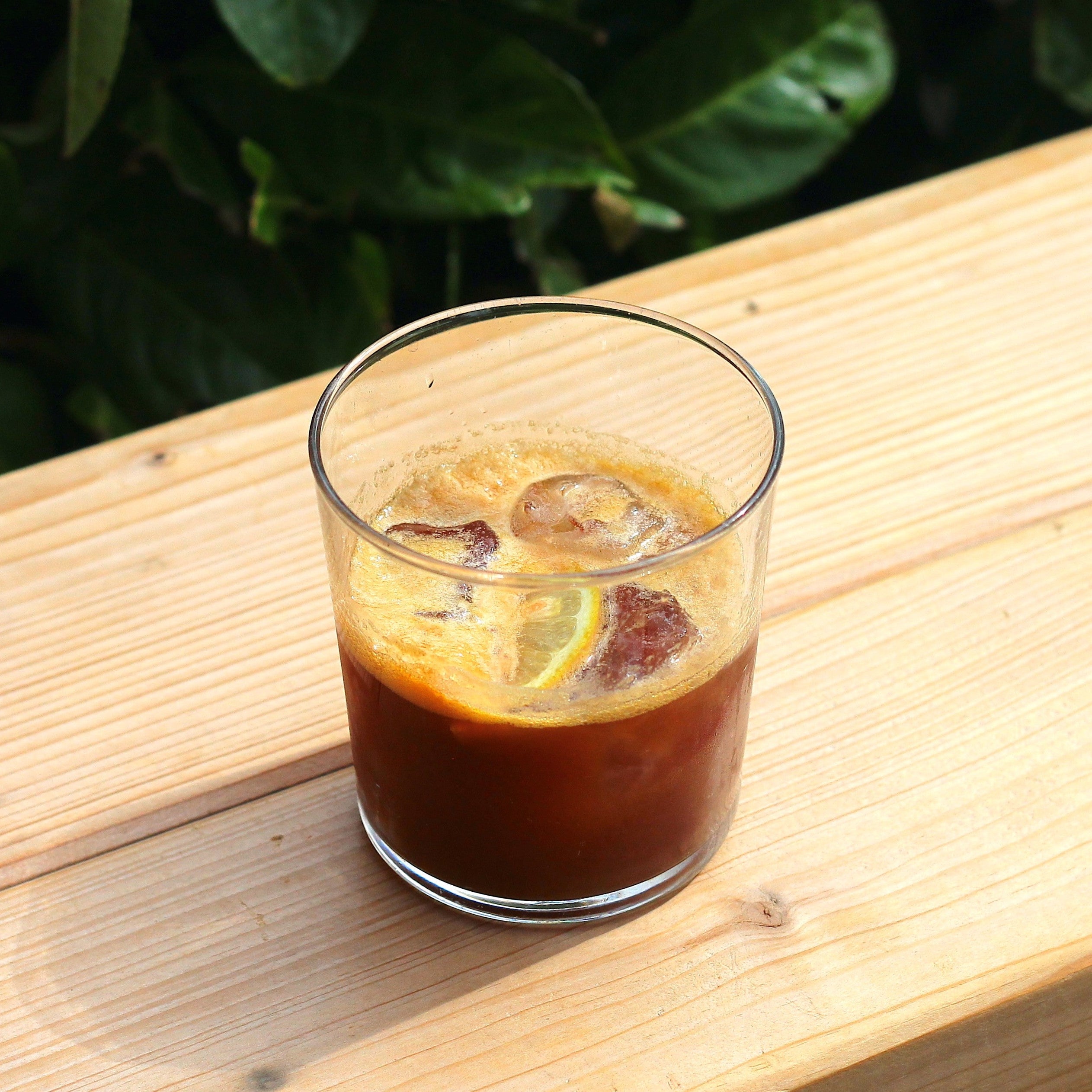 A simple glass filled with espresso tonic on the rocks garnished with a slice of fresh lemon placed on a wooden bench. A bush can be seen in the backround.