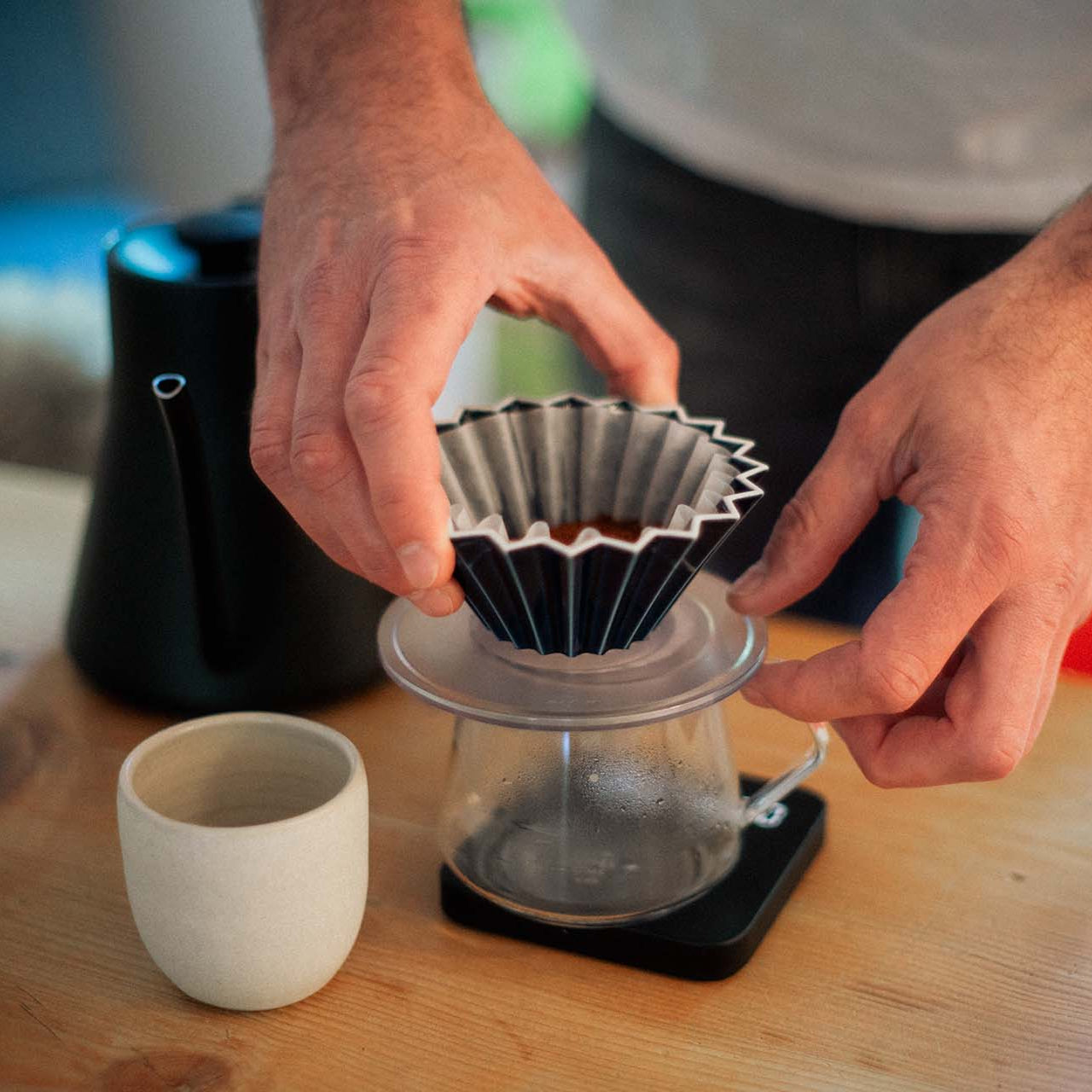 A man is carefully placing an origami dripper on a glas coffee jug that is standing on a scale.