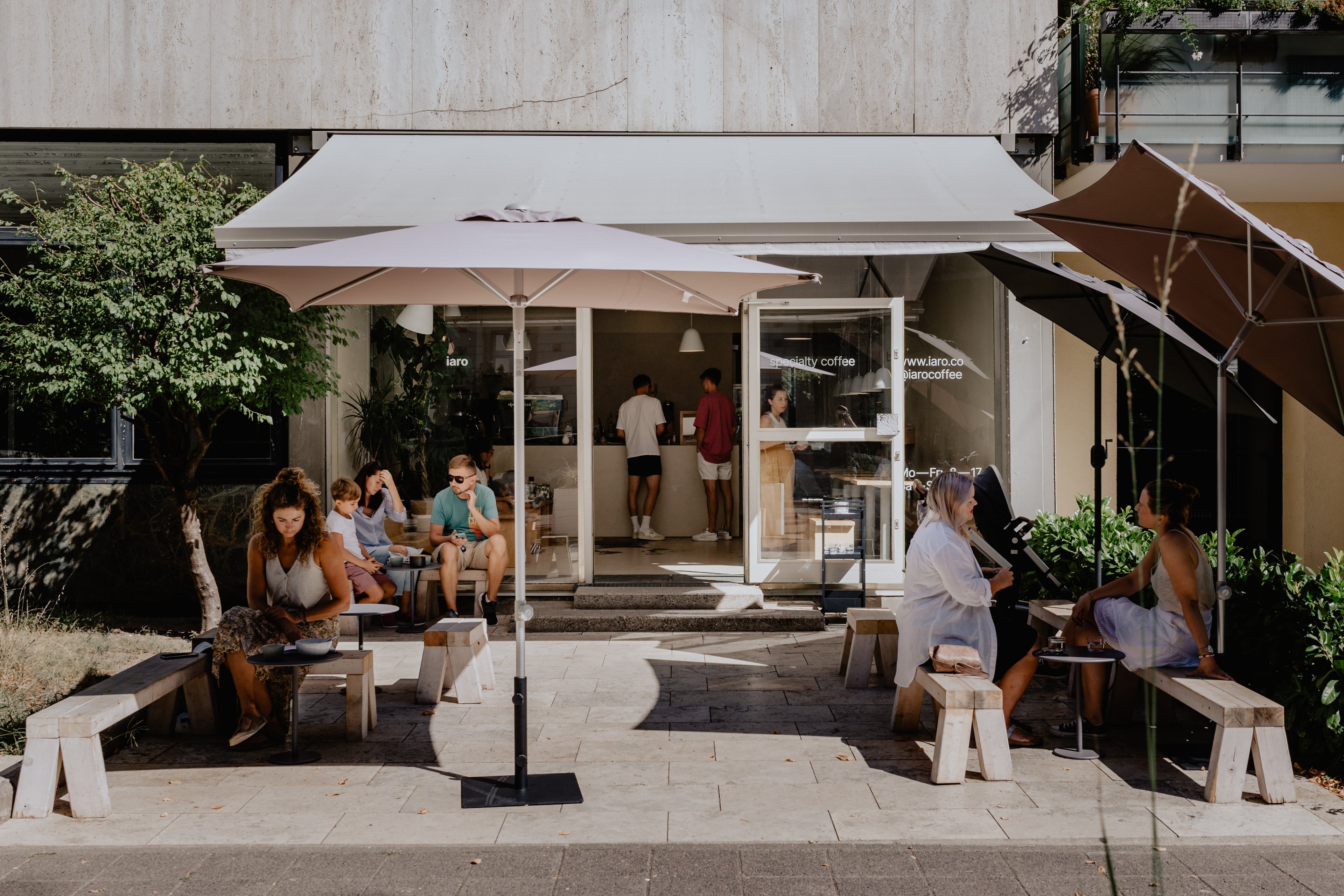 Terrace of the iaro coffeeshop in the Weststadt of Karlsruhe on a bright, sunny, summer day with people sitting on wooden stools an benches under sunshades