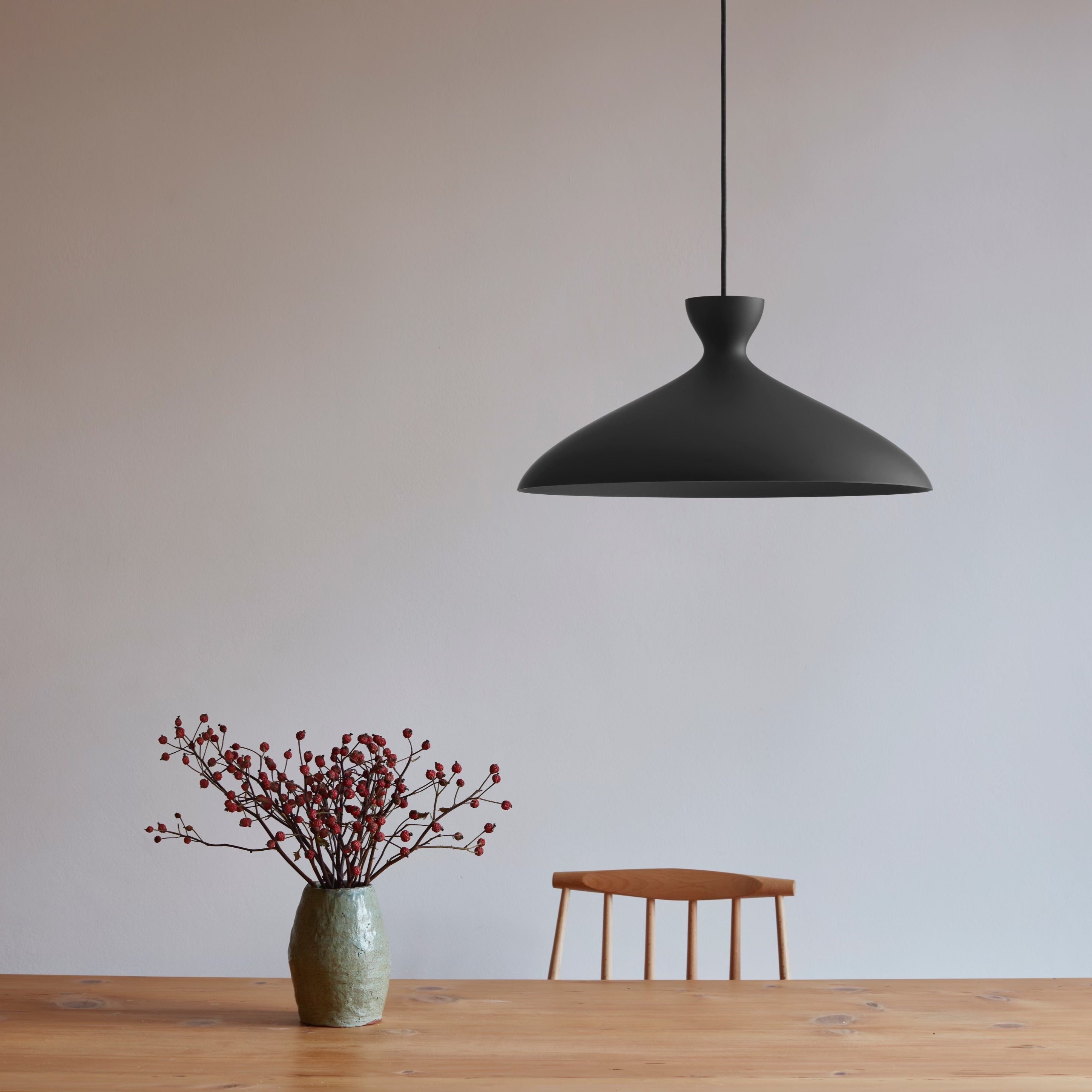 A minimalist scene with a ceramic vase on a wooden table as well as a chair behind the table. In the upper half of the fram there is a black lamp shade by nyta lighting.