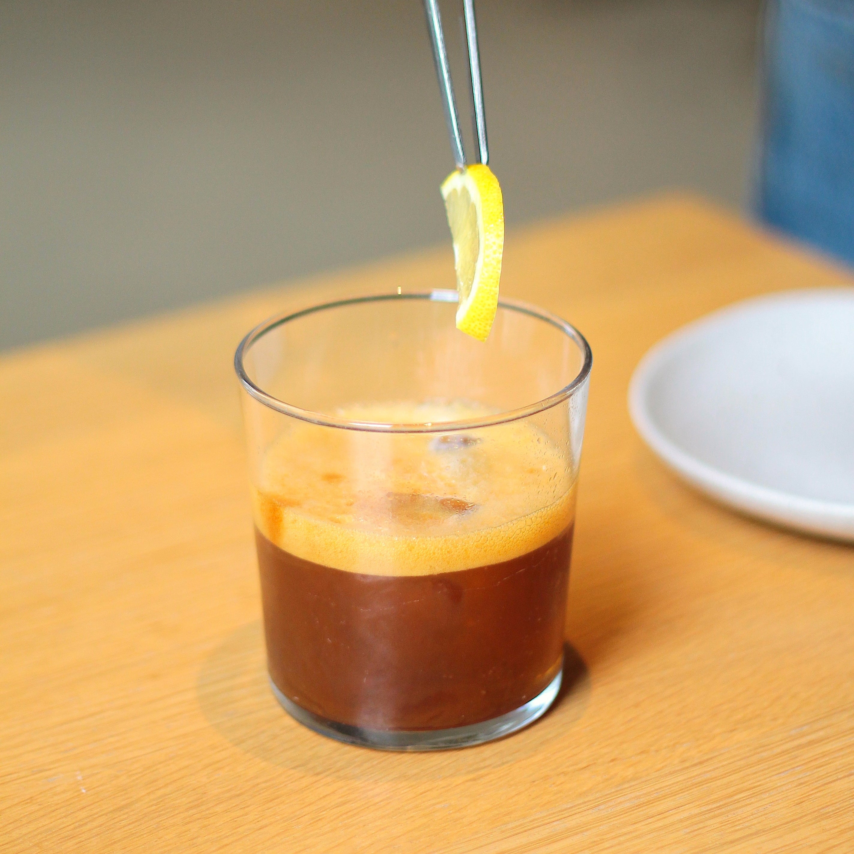 A thin slice of fresh lemon is carefully placed on top of the foam layer of an espresso tonic.