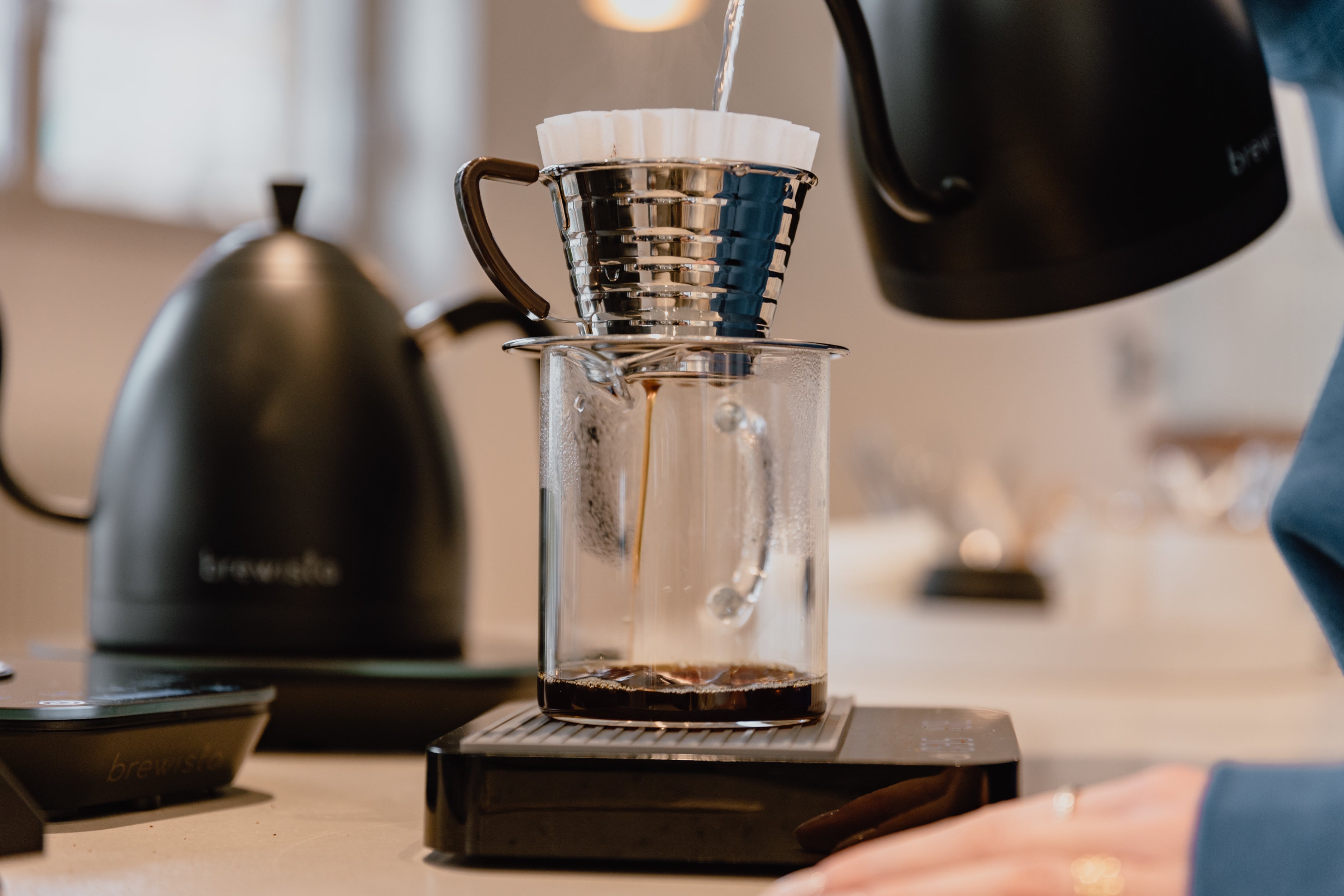 A coffee is being brewed with a kalitta dripper and filter into a delicate glass jug. The filter and jug are standing on a scale to measure the extraction.